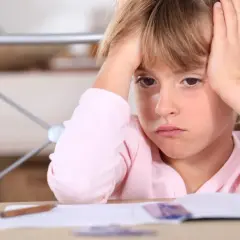 Zmartwiona dziewczynka z rękami na głowie, przy biurku z zeszytem. Czy to oznaka, że ferrytyna norma u dzieci jest nieprawidłowa?