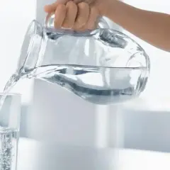 Dłoń przelewa czystą wodę alkaliczną z dzbanka do szklanki, tworząc bąbelki.