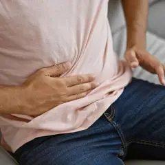 Mężczyzna siedzi na kanapie, trzymając się za brzuch. Jego dłoń uciska miejsce, gdzie odczuwa ból jelit.