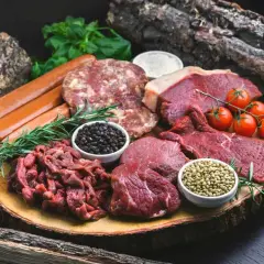Mięsne bogactwo na drewnianej desce: steki, kiełbaski, mielone, pomidorki i przyprawy. Idealne składniki dla diety carnivore.