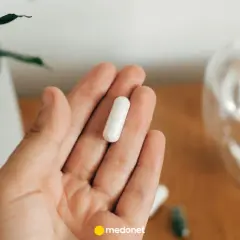 Dłoń trzyma białą kapsułkę. Obok szklanka wody i roślina. Uważaj na nadmiar magnezu objawy.