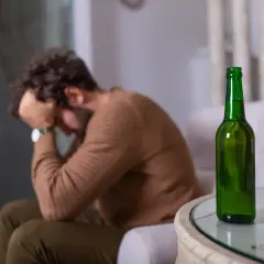 Mężczyzna siedzi ze złożonymi rękami na głowie, obok stoi butelka piwa. Może to symbolizować nagłą nietolerancję alkoholu i związane z nią problemy.