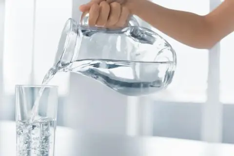 Dłoń przelewa czystą wodę alkaliczną z dzbanka do szklanki, tworząc bąbelki.