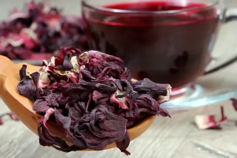 Suszone płatki hibiskusa na drewnianej łyżce i kubek z naparem. Hibiskus co to? To piękny, czerwony napój.