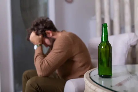 Mężczyzna siedzi ze złożonymi rękami na głowie, obok stoi butelka piwa. Może to symbolizować nagłą nietolerancję alkoholu i związane z nią problemy.