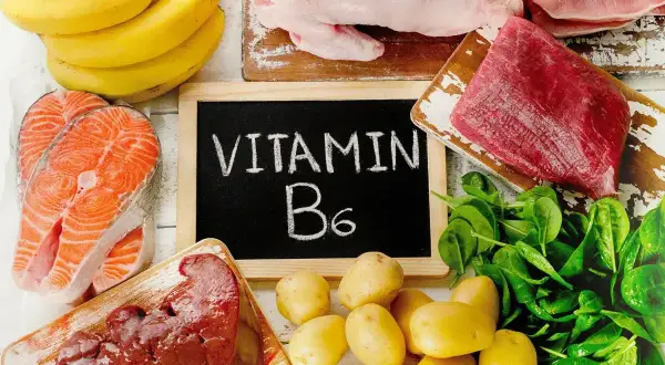 Tablica z napisem "WITAMINA B6" otoczona produktami bogatymi w tę witaminę: łososiem, bananami, wątróbką, ziemniakami, szpinakiem i mięsem.