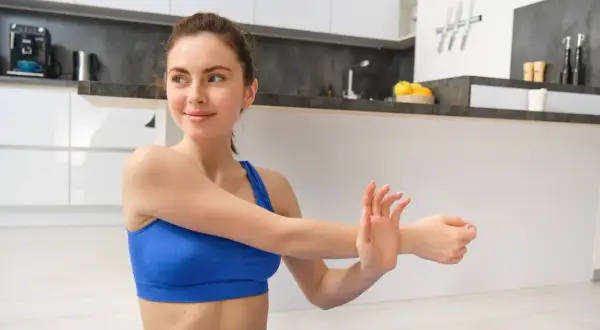 Kobieta w niebieskim sportowym topie rozciąga rękę, próbując złagodzić bolesne skurcze mięśni po treningu w kuchni.