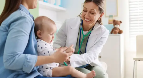Uśmiechnięta lekarka bada niemowlę, które siedzi na kolanach mamy. Wykonuje test na odwodnienie, delikatnie dotykając rączki dziecka.