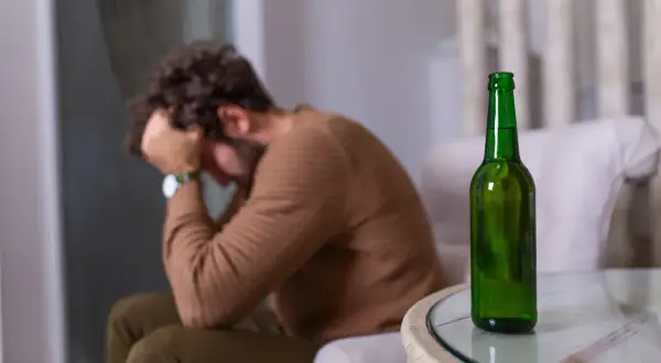 Mężczyzna siedzi ze złożonymi rękami na głowie, obok stoi butelka piwa. Może to symbolizować nagłą nietolerancję alkoholu i związane z nią problemy.