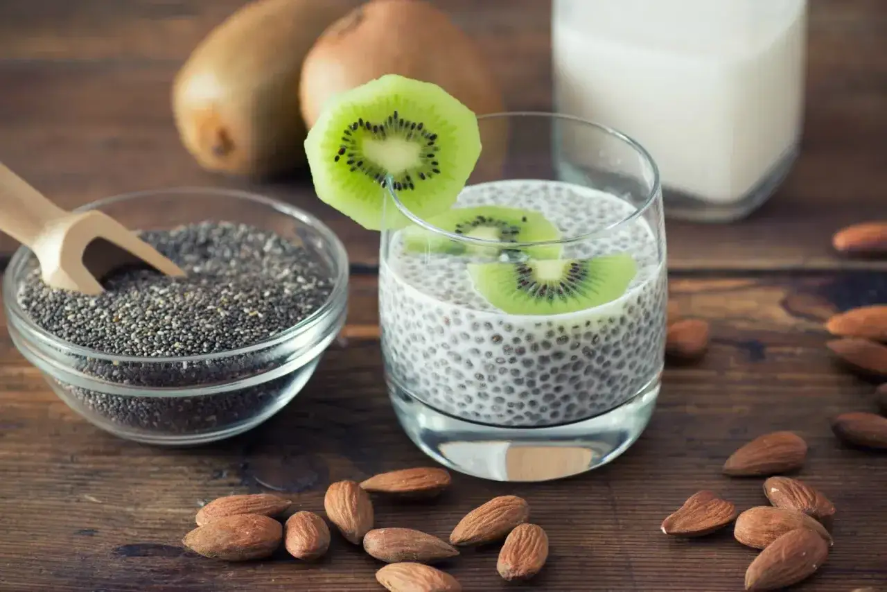 Pudding z nasion chia z kiwi i migdałami. Obok miseczka z nasionami chia i mleko.