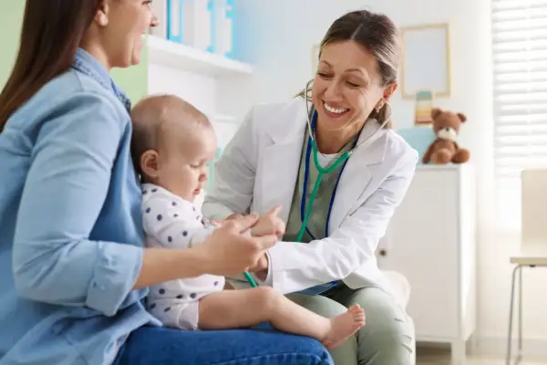 Uśmiechnięta lekarka bada niemowlę, które siedzi na kolanach mamy. Wykonuje test na odwodnienie, delikatnie dotykając rączki dziecka.